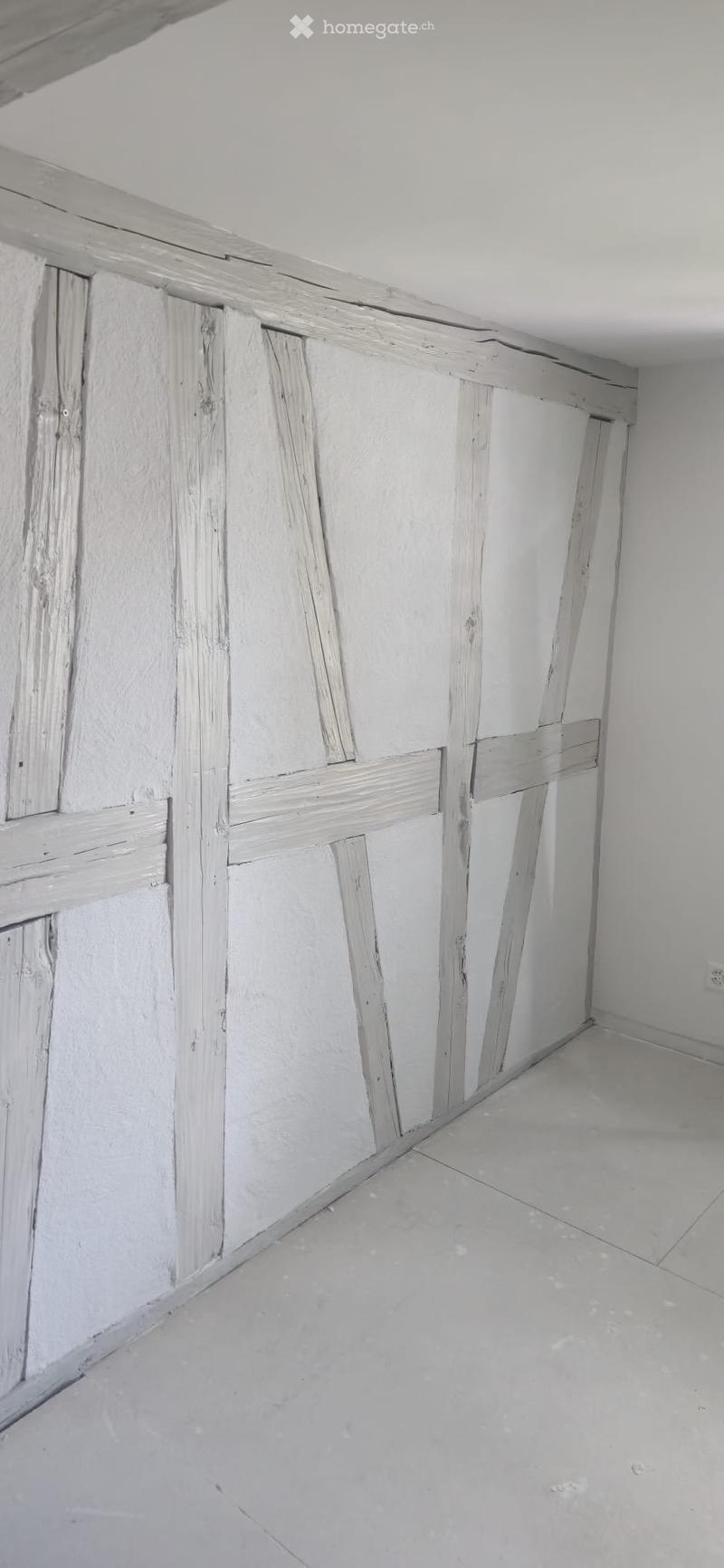 Empty room with exposed wooden paneling on one wall and a smooth white wall on the other. The floor is a light grey concrete or tile surface.