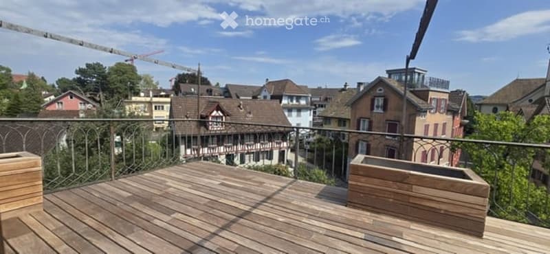 Wooden balcony with metal railing, overlooking a residential area with traditional houses and greenery.