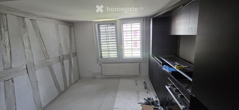 Empty room with unfinished walls, tiled floor, and a window with blinds. Kitchen cabinets are visible on one side.