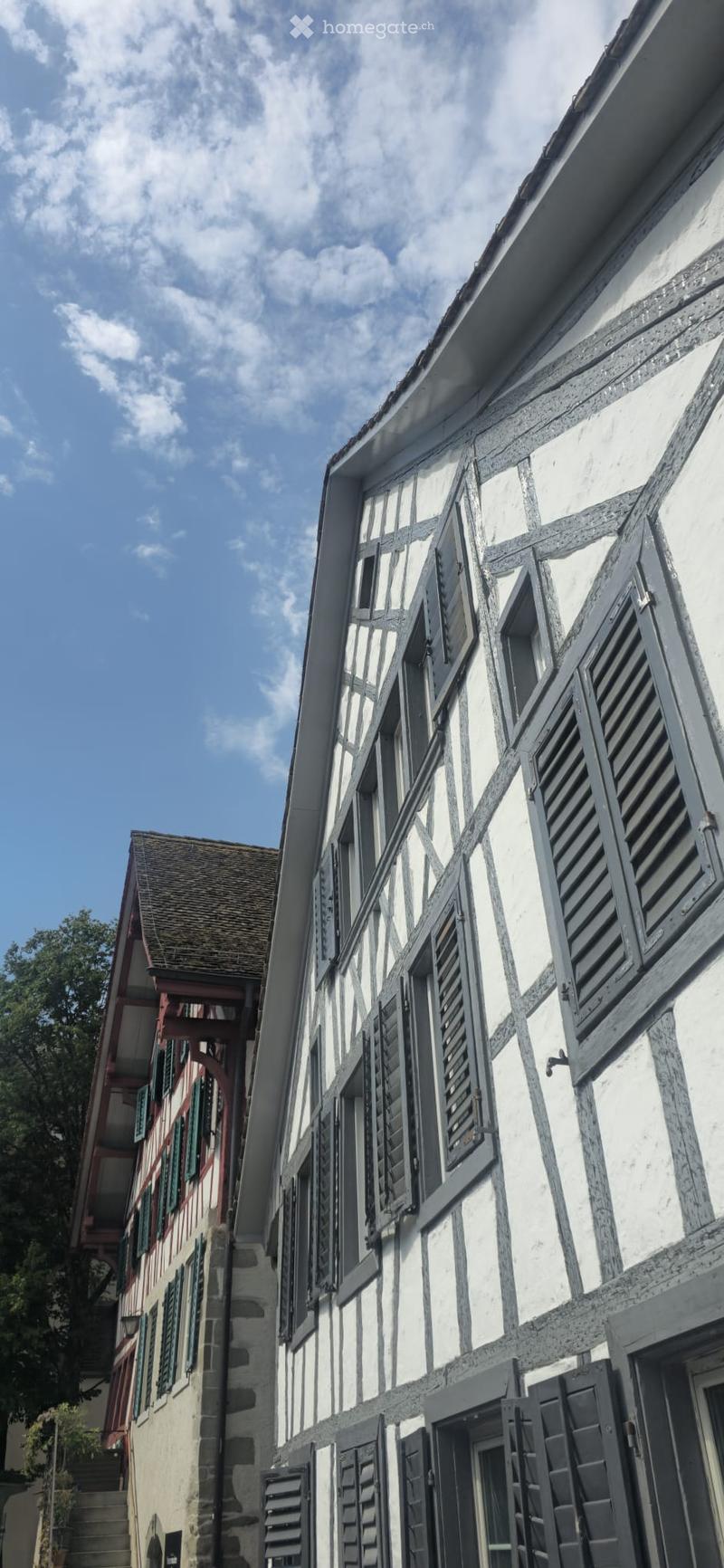 A two-story half-timbered building with white plaster and dark wooden beams, featuring multiple windows with shutters and a visible doorway.