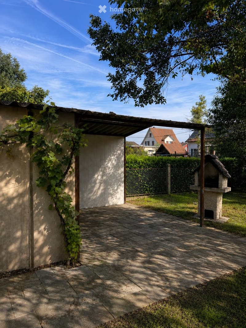A covered outdoor area with a brick barbecue, wooden pergola, and surrounding greenery.