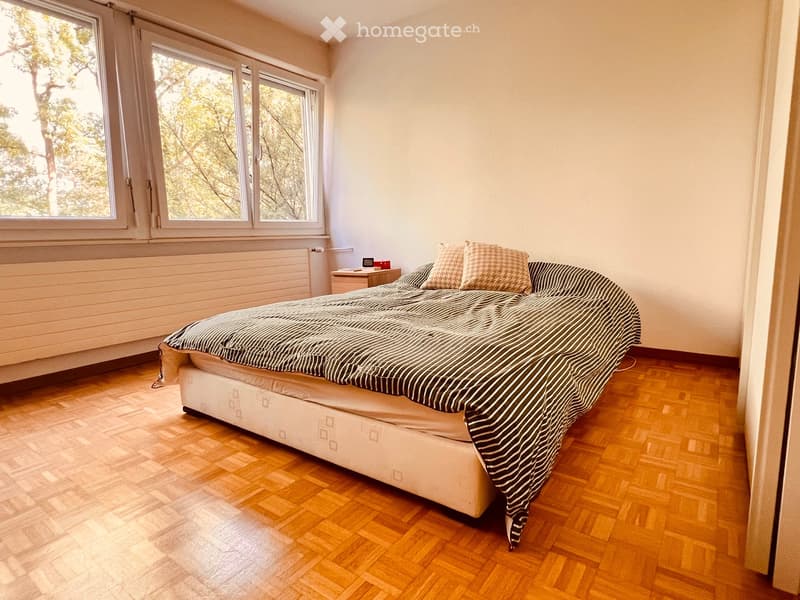 Bright bedroom with wooden flooring, large windows, a neatly made bed with striped bedding, a nightstand, and a radiator below the windows.