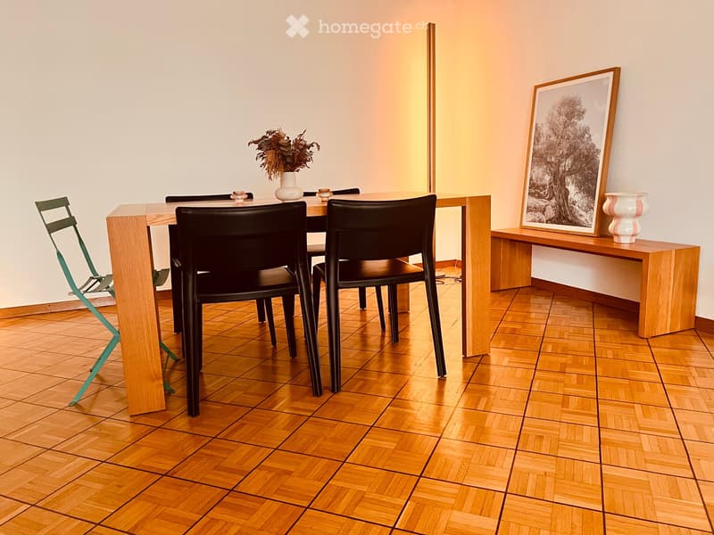 Dining area with a wooden table, black chairs, a vase with dried flowers, a picture frame on a shelf, and a green folding chair.