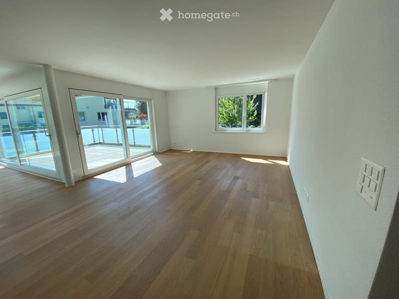 Empty room with light wooden flooring, white walls, large sliding glass doors leading to a balcony, and a window with a view of greenery.
