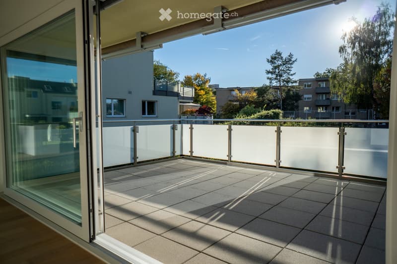 Modern balcony with tiled floor, glass sliding doors, and a metal railing, overlooking a residential area with trees and buildings.