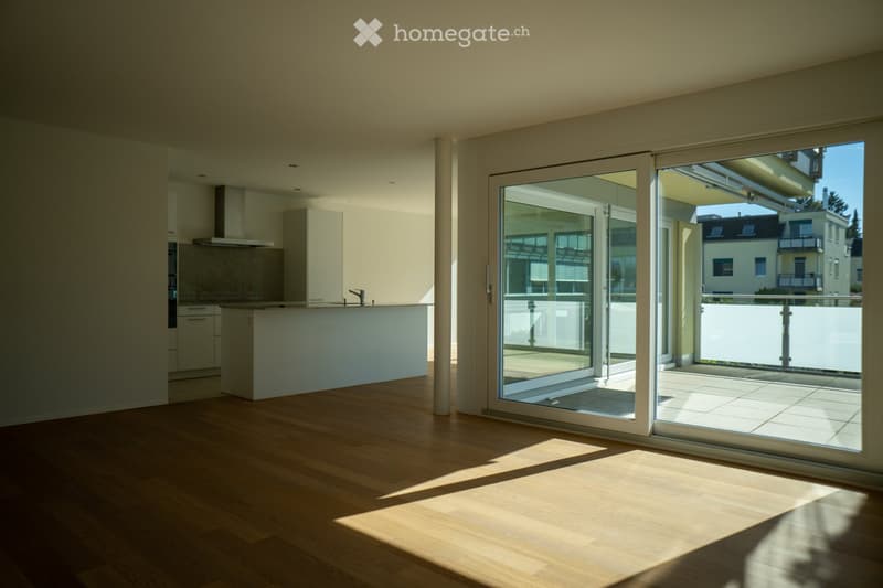 Bright, modern kitchen with light wooden flooring, white cabinetry, and a central island. Large sliding glass doors open to a balcony with a view of neighboring buildings.
