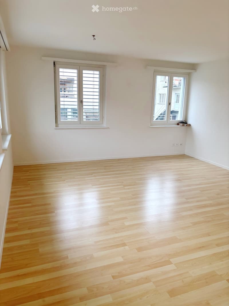 Empty room with light wooden flooring, white walls, and large windows with blinds. No furniture or equipment present.