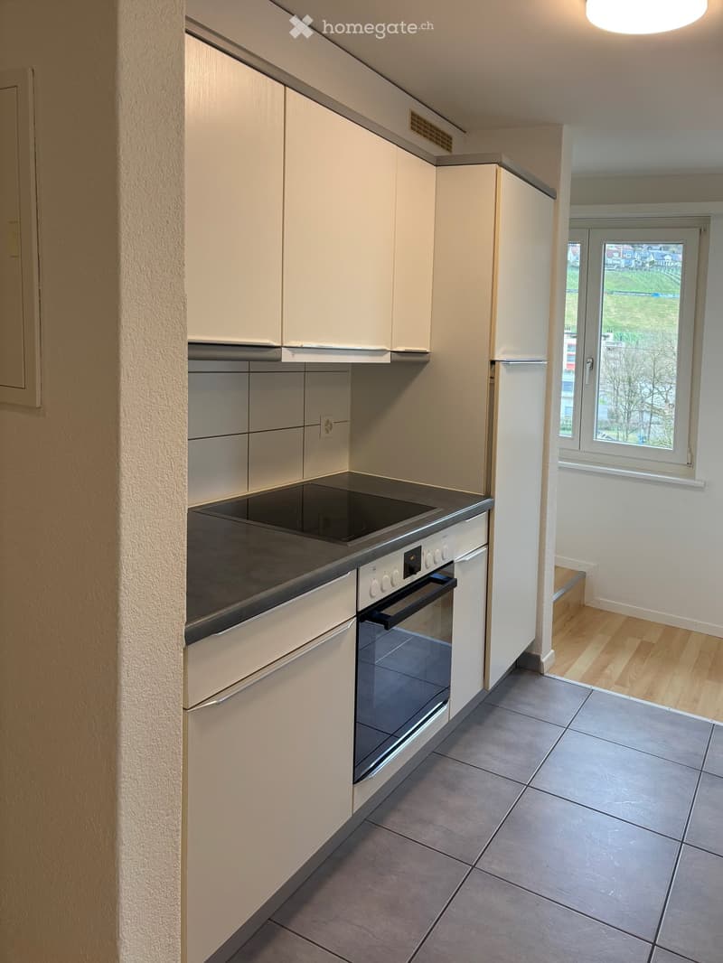 Modern kitchen with white cabinets, an induction cooktop, and an oven. The space features tiled flooring and a window offering a view of the outdoors.