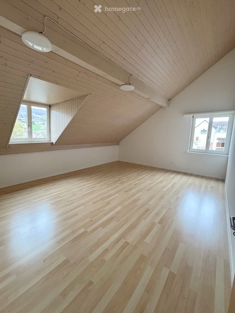 Empty attic room with wooden floors, white walls, and exposed wooden ceiling beams. Two windows provide natural light.