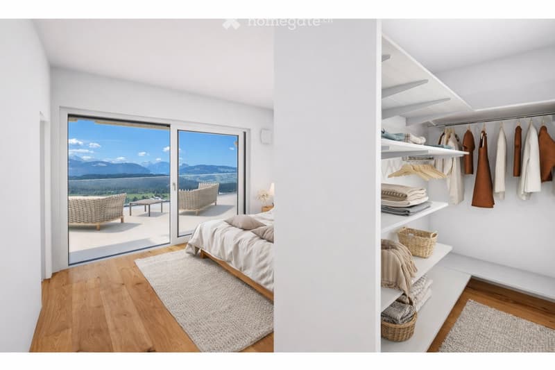 Bright bedroom with wooden flooring, large glass doors opening to a balcony with outdoor seating and scenic mountain view. Includes a neatly made bed, a small table, and a built-in wardrobe with shelves and hanging space.