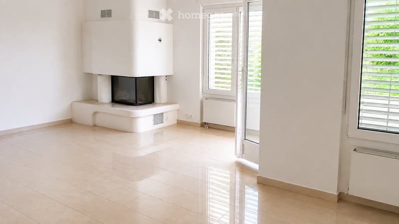 Empty room with tiled floor, white walls, fireplace, and large windows with blinds.