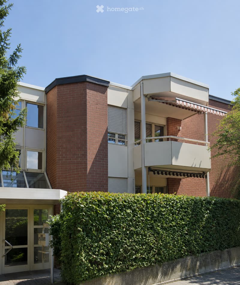 Modern two-story building with red brick and white panels, featuring large windows, a balcony with an awning, and a well-maintained hedge in front.