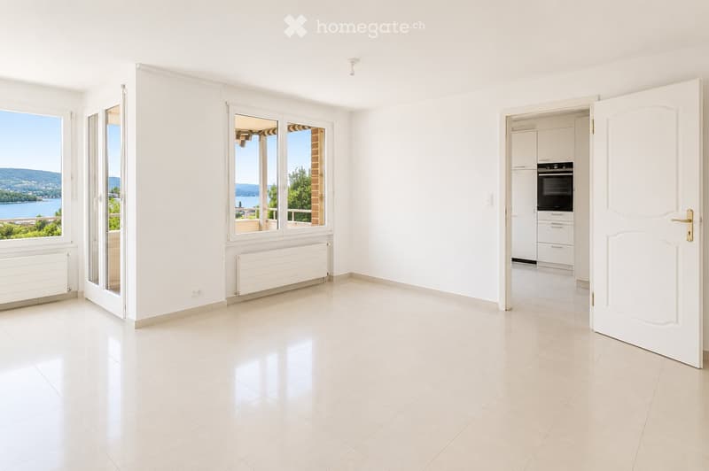 Bright, empty room with white tiled flooring, large windows offering scenic views, and a door leading to a kitchen area.