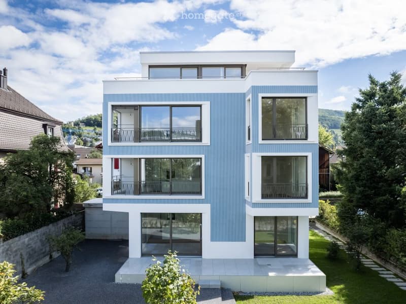Modern two-story house with blue and white exterior, large windows, balconies, and a raised ground floor.