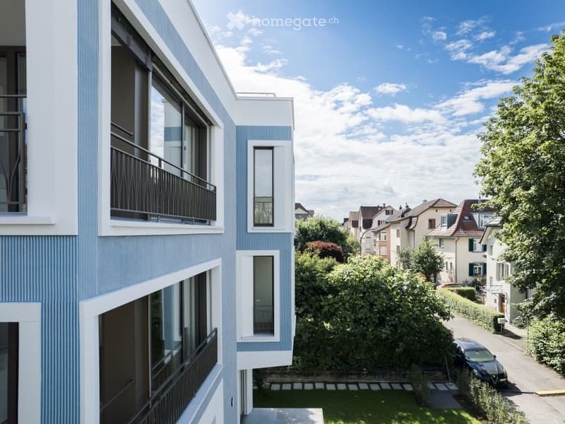 Modern multi-story building with large windows and balconies, surrounded by greenery and neighboring houses.