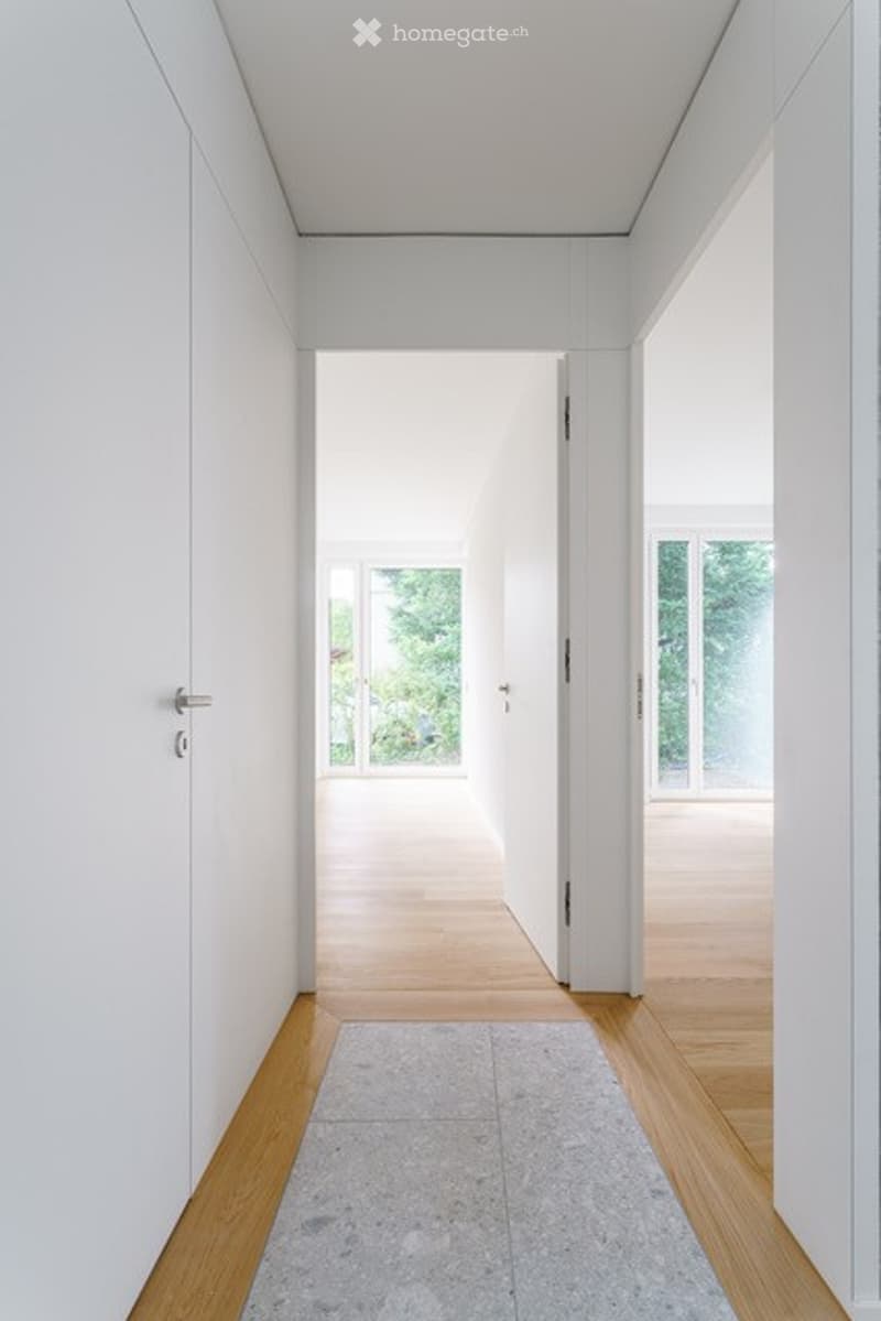 Minimalist hallway with white walls, wooden flooring, and large windows at the end, leading to an open space.
