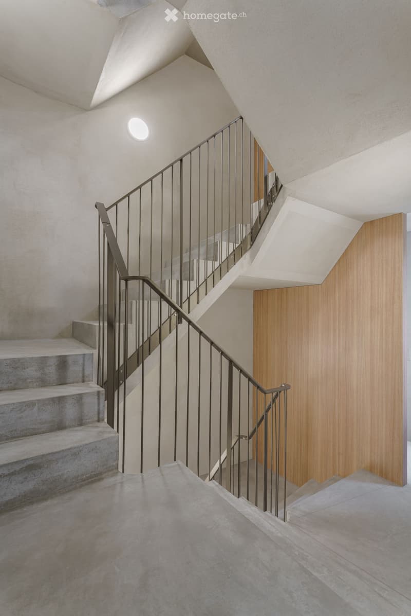 Modern staircase with concrete steps, sleek metal railing, and warm wooden paneling on one wall. The ceiling features recessed lighting and a geometric design.
