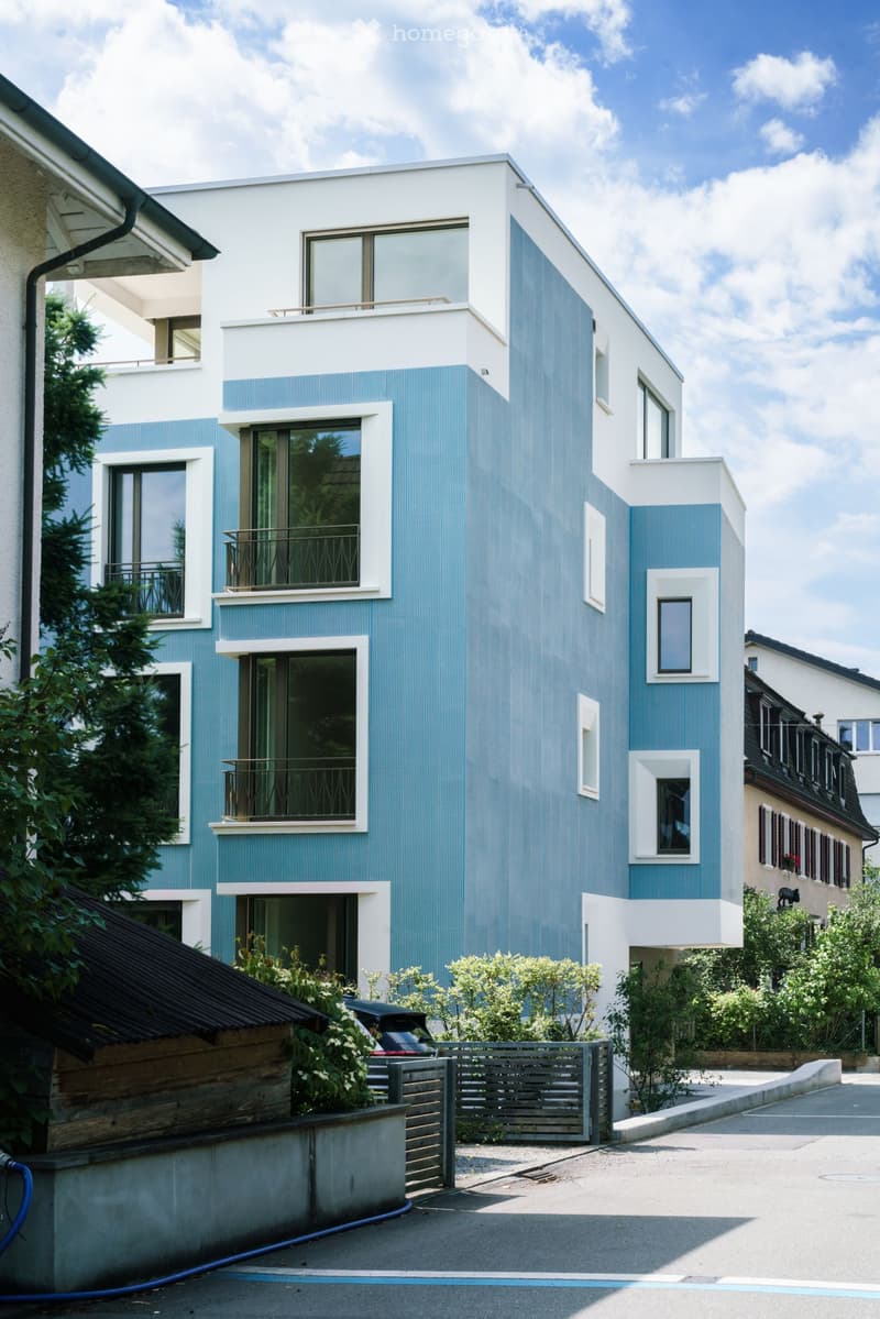 Modern two-story house with blue and white exterior, large windows, balconies, and a fenced garden area.