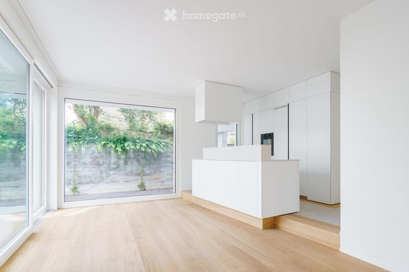 Modern, minimalist kitchen with white cabinets, a central island, and large windows overlooking a garden.