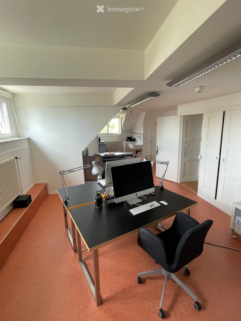 Modern office space with a black desk, ergonomic chair, computer setup, and built-in shelving. Features large windows, warm-toned carpet, and a small adjacent bathroom area.