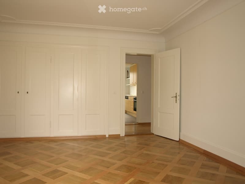 Empty room with wooden parquet flooring, white walls, and built-in white closet units. An open doorway leads to a kitchen area with light-colored cabinets and appliances.
