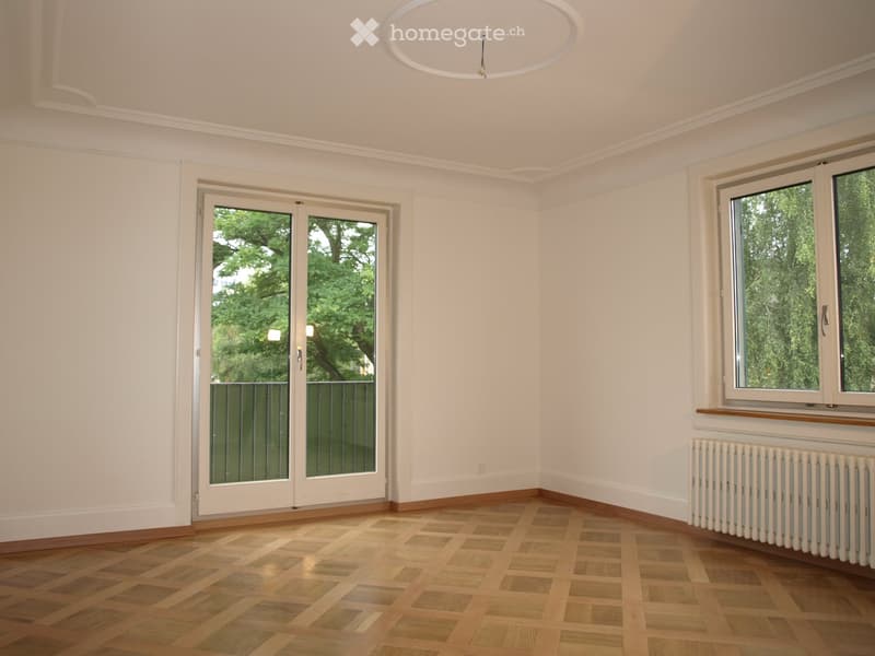 Empty room with white walls, wooden parquet flooring, large windows, and a sliding glass door leading to a balcony.