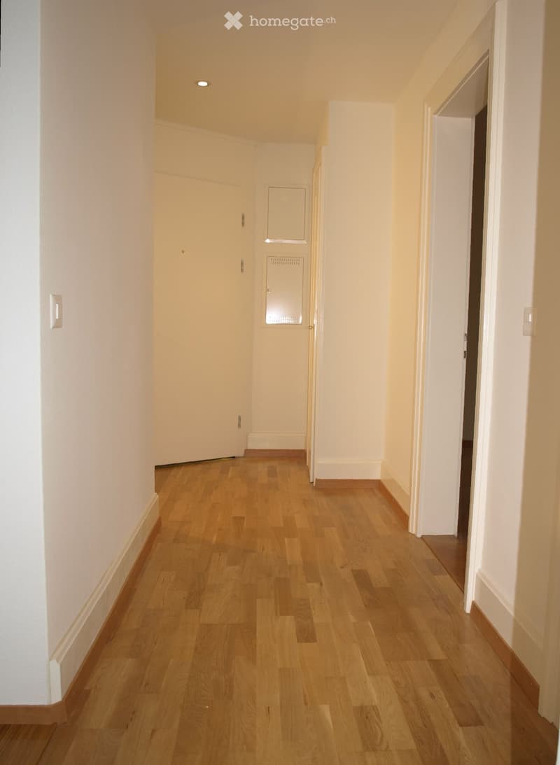 A narrow hallway with light wooden flooring and white walls, featuring a closed door on the left and an open doorway on the right leading to another room.