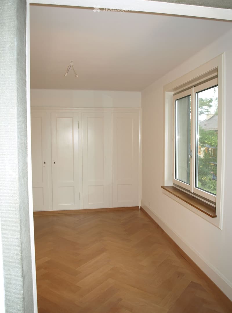 Empty room with wooden herringbone flooring, white walls, built-in white cabinetry, large windows with wooden sills, and a hanging light fixture.