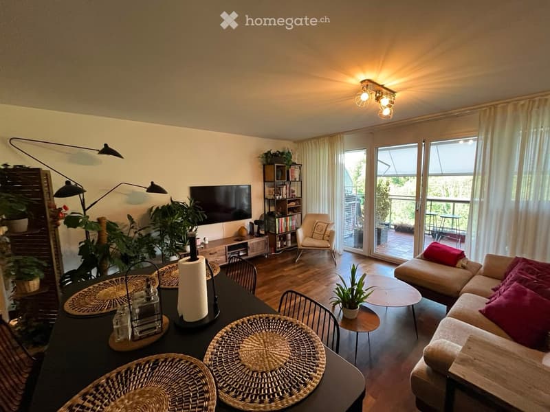 Bright living room with large windows and sliding glass doors leading to a balcony. Features a beige sofa with red cushions, a coffee table, a dining table with chairs, a television, a bookshelf, and several plants.
