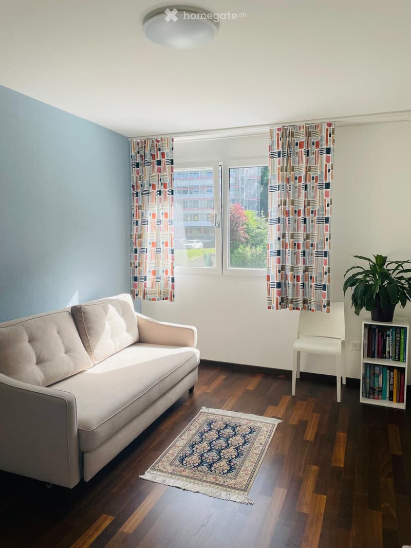 Living room with a light blue wall, wooden floor, and large windows with colorful curtains. Features a beige sofa, white chair, bookshelf with books, a potted plant, and a patterned rug.
