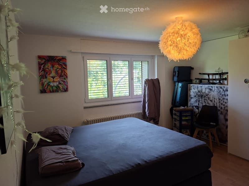 Cozy bedroom with a double bed, pillows, a hanging lamp, a chair, a small table, a painting of a lion, and a refrigerator with a decorative cover.