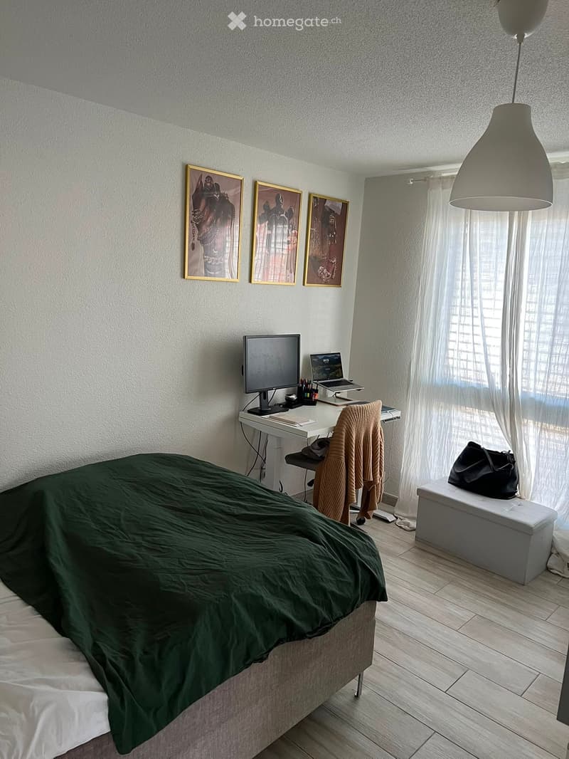 Modern bedroom with a bed covered in green duvet, a desk with a laptop and monitor, a chair, and a small white bench with a black bag on it. The room features light wooden flooring and white walls with framed artwork.