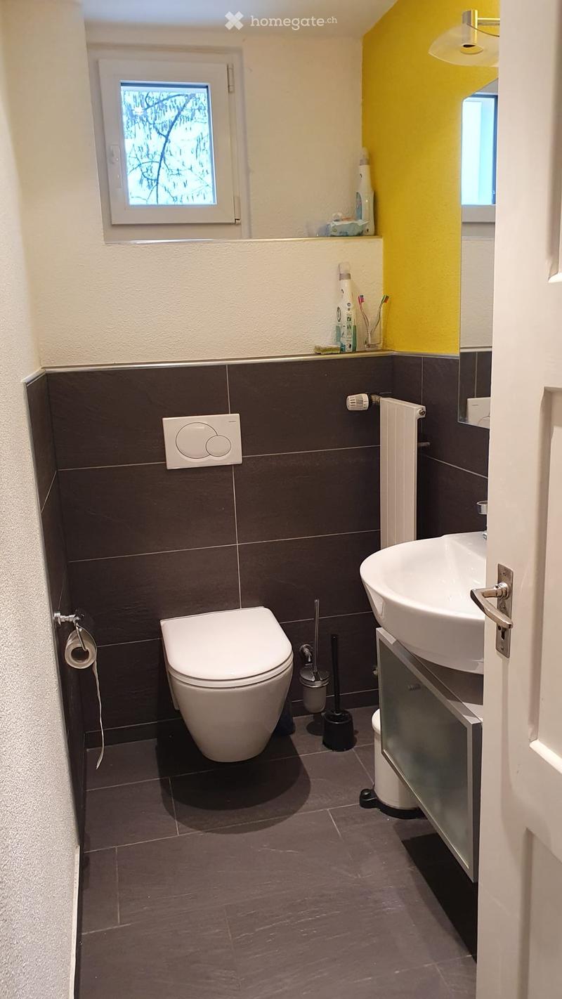 Modern bathroom with dark tiled walls and floor, white toilet, sink, and mirror, and a small window with frosted glass.