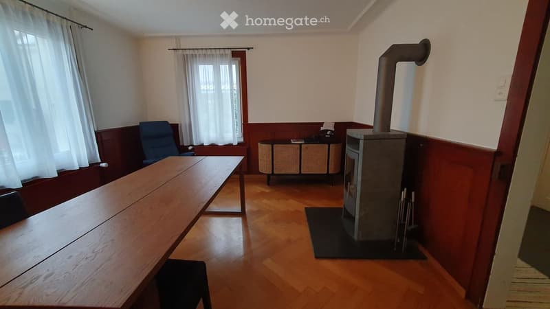 A room with wooden flooring and walls, featuring a long wooden table, a blue chair, a wooden stove, and a storage unit with wicker baskets.