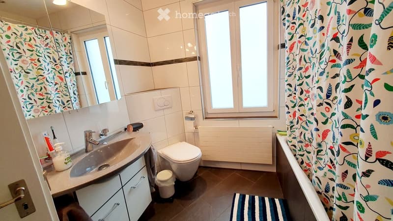 Modern bathroom with white tiles, a large window, a sink with a mirror, a toilet, and a shower curtain. Includes a soap dispenser, toothpaste, and a small trash bin.