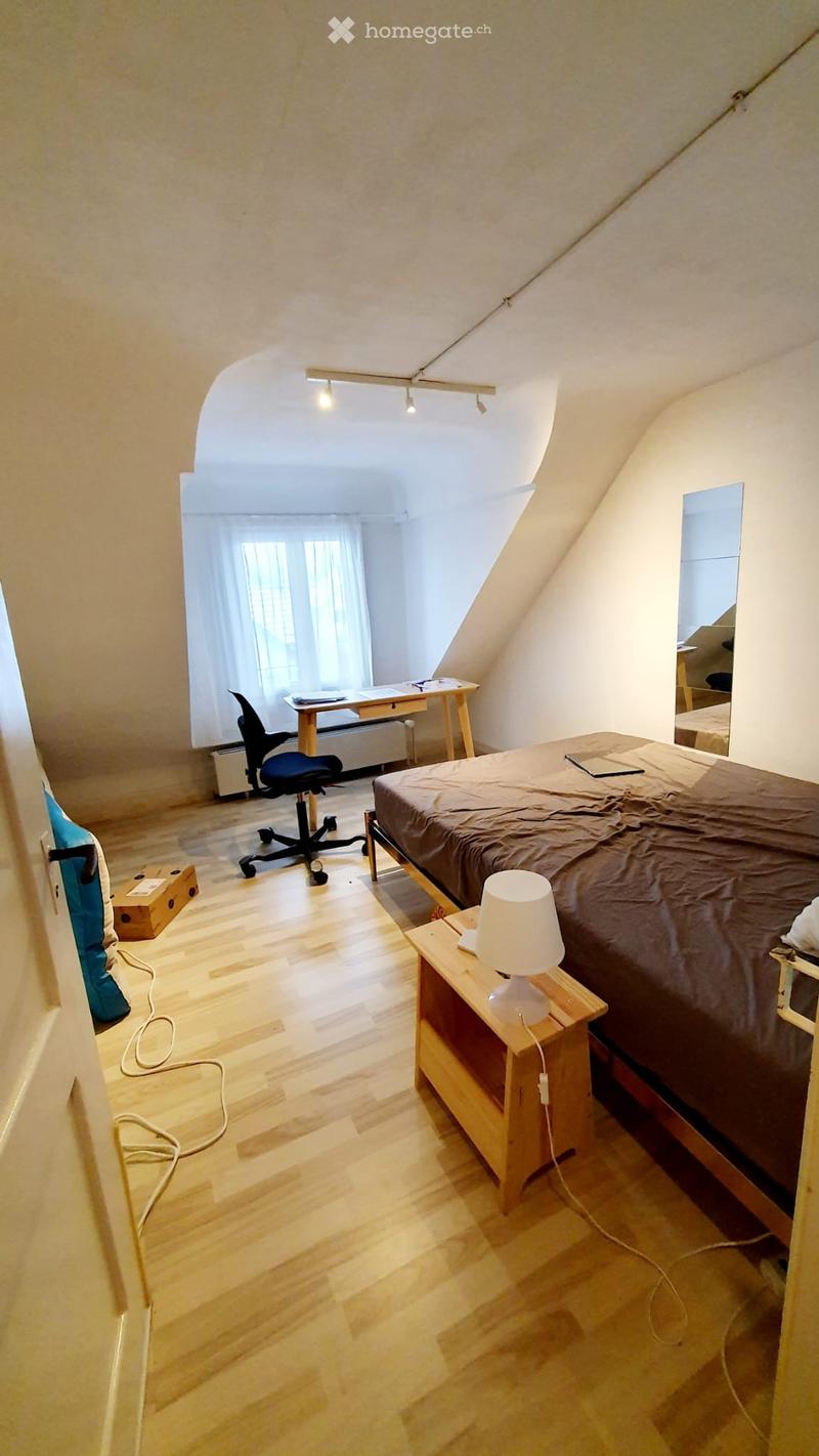 Cozy attic bedroom with wooden flooring, slanted ceilings, and ample natural light from large windows. Features a bed with a brown duvet, a nightstand with a lamp, a desk with a chair, and a mirror in the adjacent room.