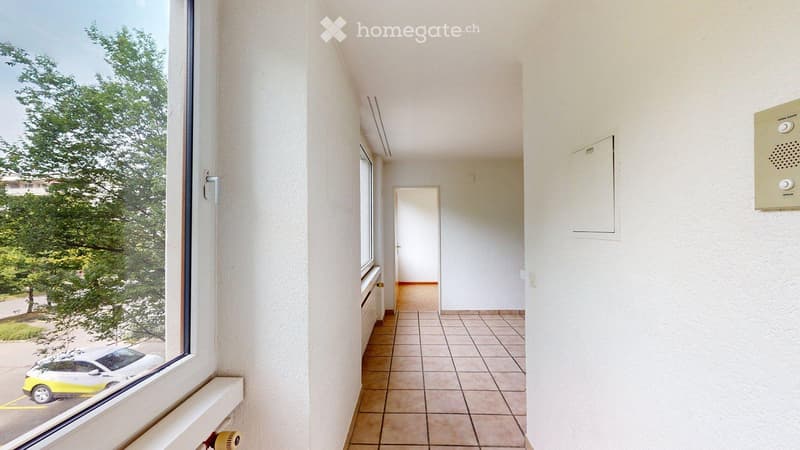 Empty hallway with tiled floor, white walls, and a window showing a view of a parked car and greenery outside.