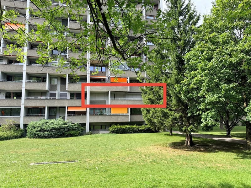 A multi-story residential building with balconies and windows, surrounded by green trees and a grassy lawn.