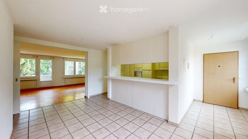 Empty room with tiled floor, wooden door, and a small kitchen area with cabinets and a countertop.