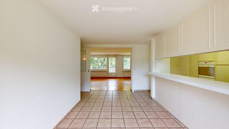 Empty hallway with tiled floor and white walls, leading to an open living space with wooden flooring and large windows.