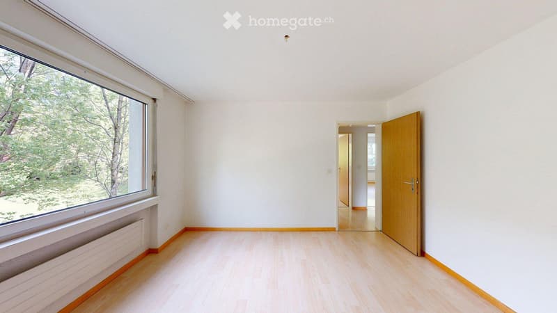 Empty room with light wooden flooring, white walls, large window with view of greenery, and a wooden door leading to another space.
