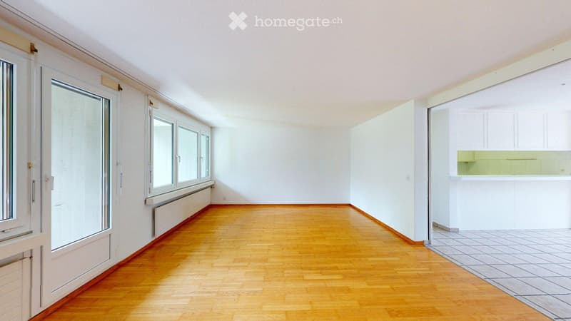 Empty room with wooden flooring, white walls, large windows, and a small kitchen area visible in the background.