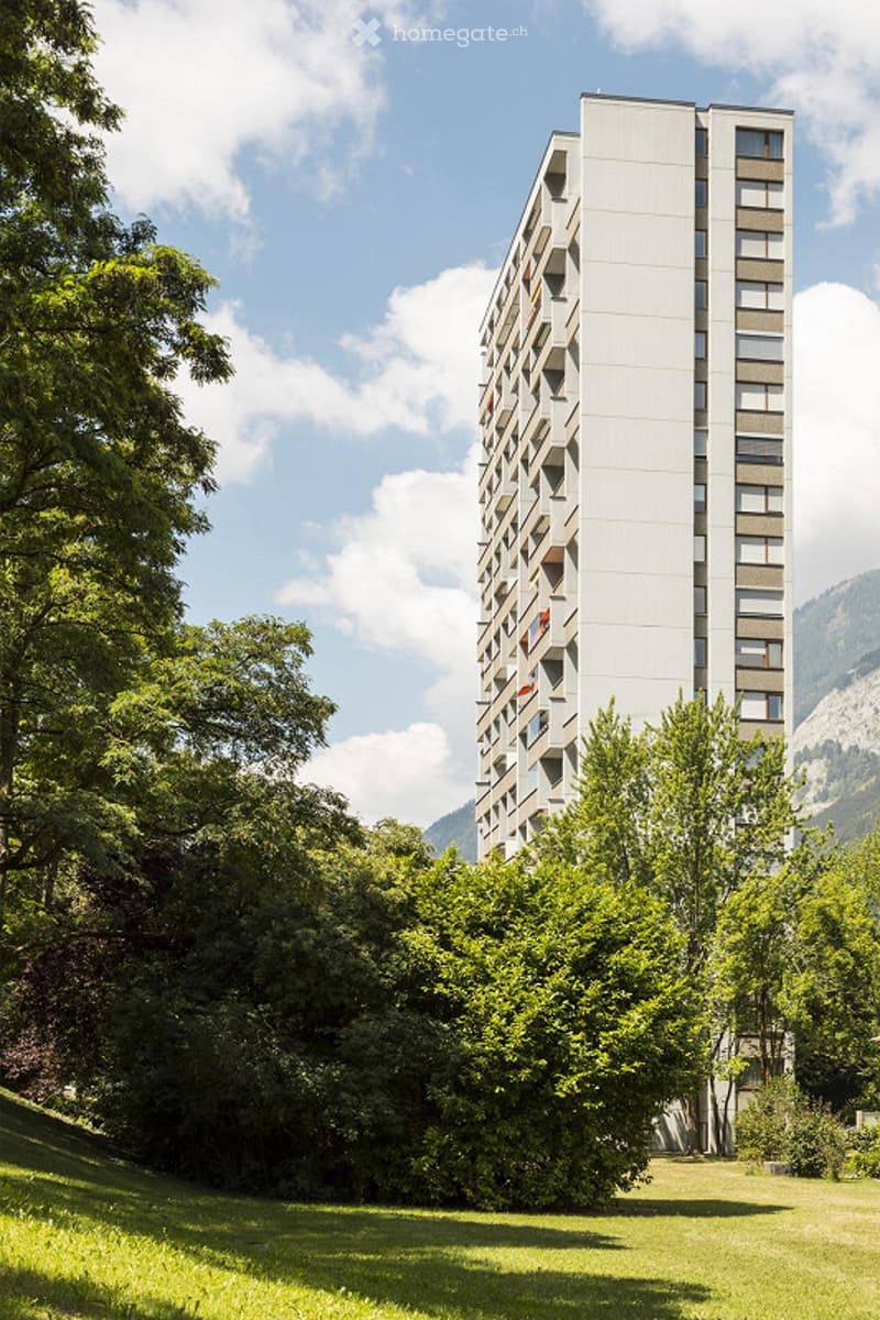 Modern high-rise apartment building with balconies, surrounded by lush green trees and a grassy lawn under a partly cloudy sky.
