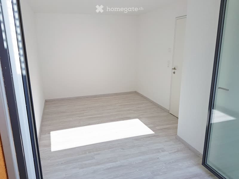 Empty room with light wooden flooring, white walls, and a white door visible in the background.
