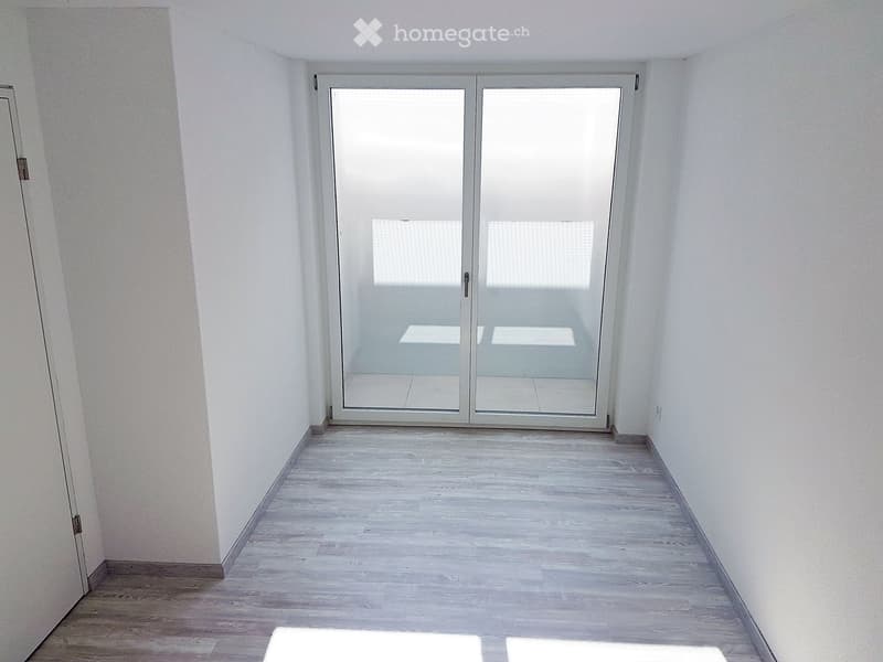Empty room with light wooden flooring and white walls, featuring a large glass sliding door leading outside.