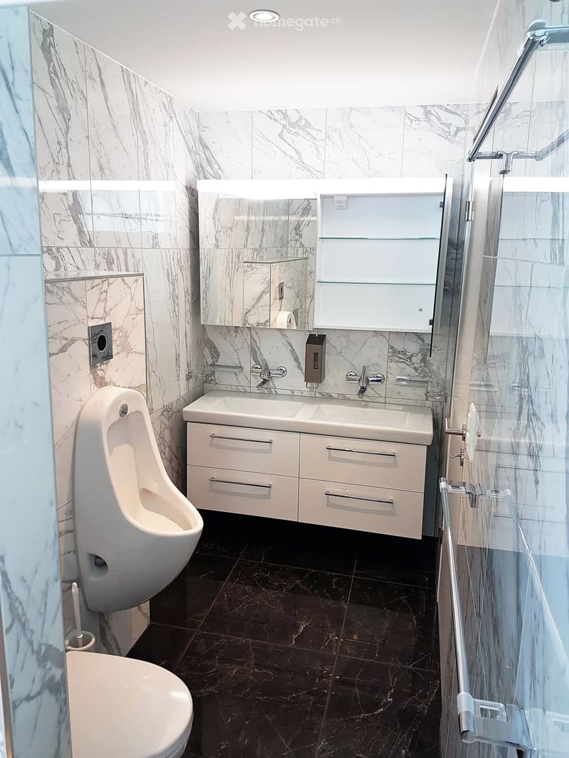 A bathroom with marble flooring and walls, a white vanity with double sinks, a urinal, a shower area with a glass door, and a toilet. It has a contemporary and minimalist design.