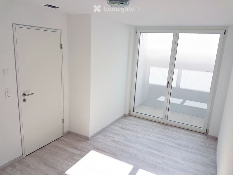 Empty room with light wooden flooring, white walls, a closed door, and a large sliding glass door leading outside.