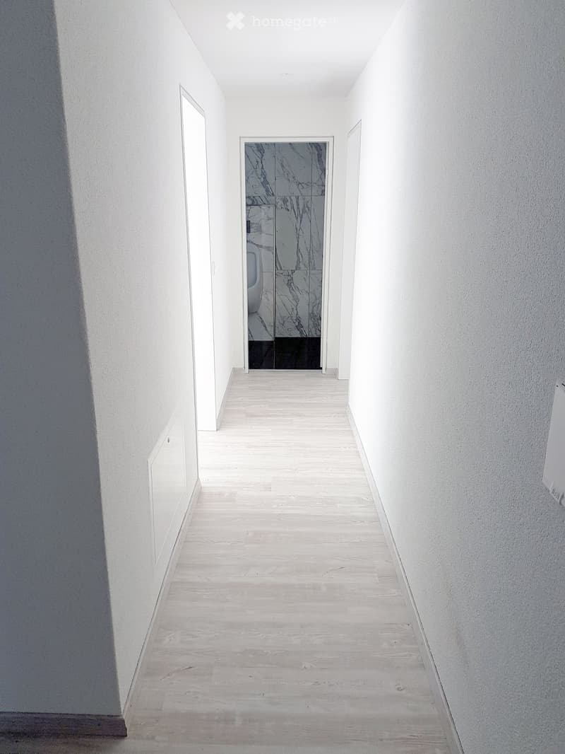 Empty hallway, white walls, wooden flooring, two doors, one leading to a bathroom