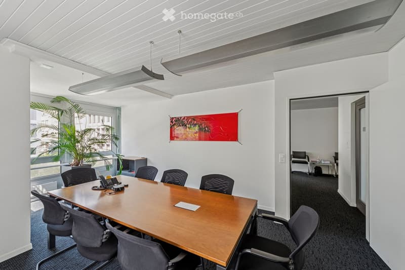 Modern office meeting room with a large wooden table, several chairs, a telephone, a potted plant, and a colorful abstract artwork on the wall. Large windows provide natural light.