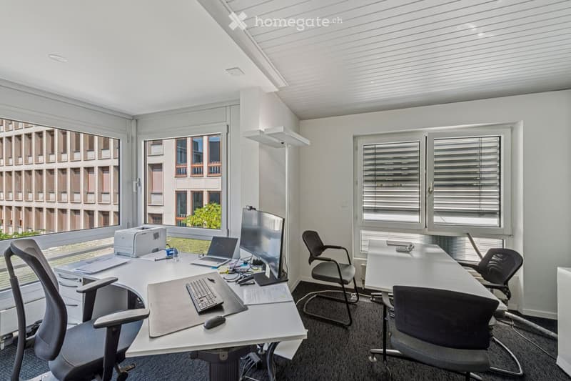 Modern office space with large windows, two desks, multiple chairs, laptops, a printer, and computer peripherals.